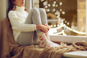 PTSD, sadness and people concept - sad beautiful girl in sweater sitting on sill at home window over christmas lights background